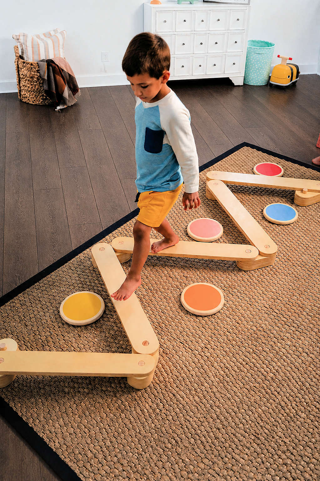 A young boy in a blue and white shirt walks barefoot across a colorful wooden balance beam and stepping stones set, placed on a textured rug in a bright, airy.