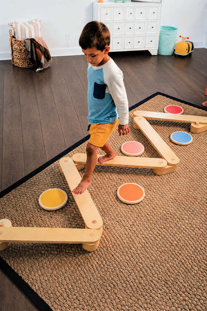 A young boy in a blue and white shirt walks barefoot across a colorful wooden balance beam and stepping stones set, placed on a textured rug in a bright, airy.