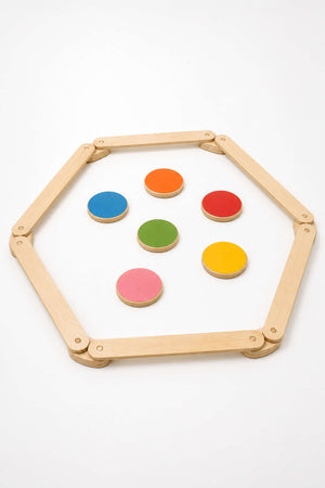 A hexagon-shaped wooden frame surrounds colorful, circular stepping stones in vibrant red, green, pink, yellow, and blue. This sustainable play set encourages creative movement and coordination for children.