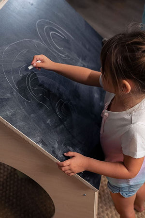 A young girl draws with chalk on the blackboard roof of a wooden playhouse, showcasing its interactive design. The pastel-colored playhouse features natural wood, promoting imaginative play for children.