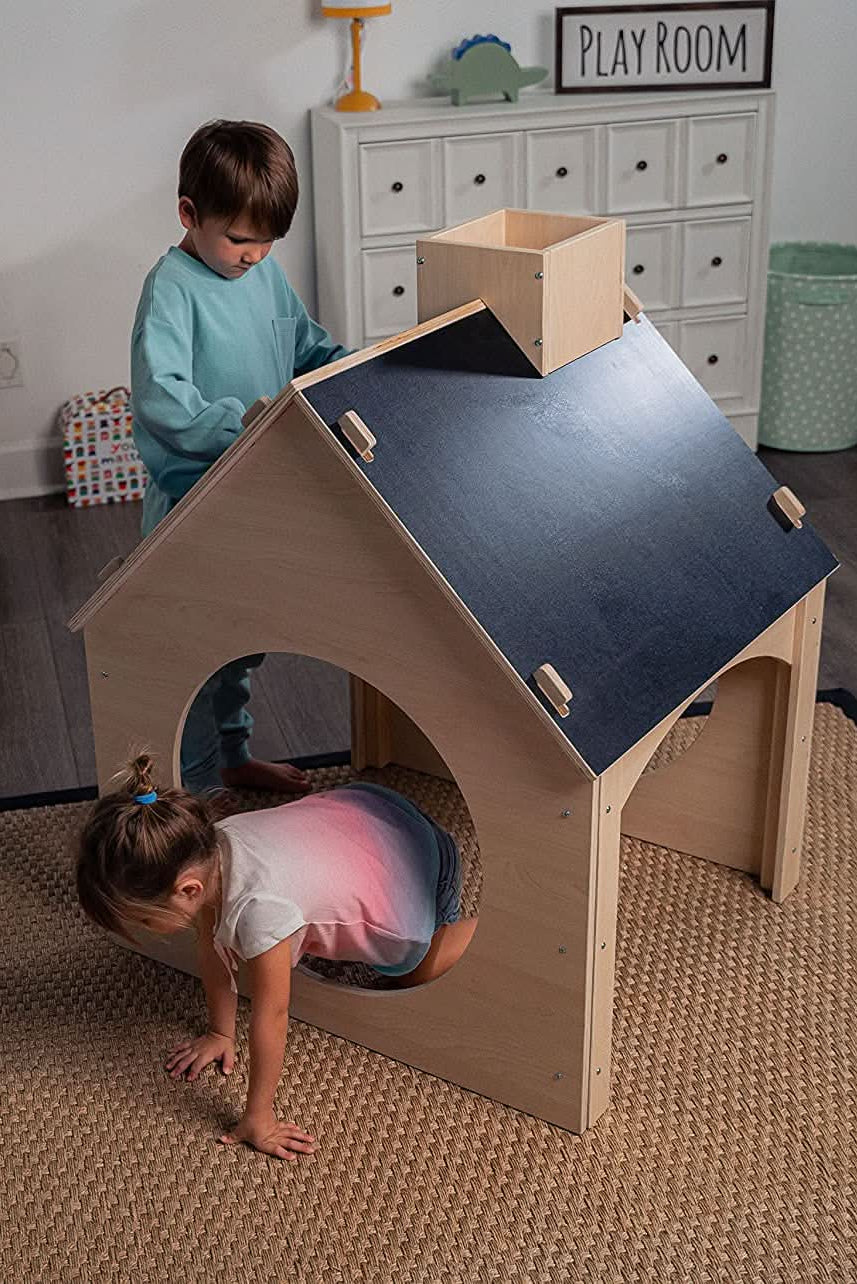 Two children play in a wooden childrens playhouse featuring a chalkboard roof. The playhouse is set in a cozy playroom with a natural fiber rug.