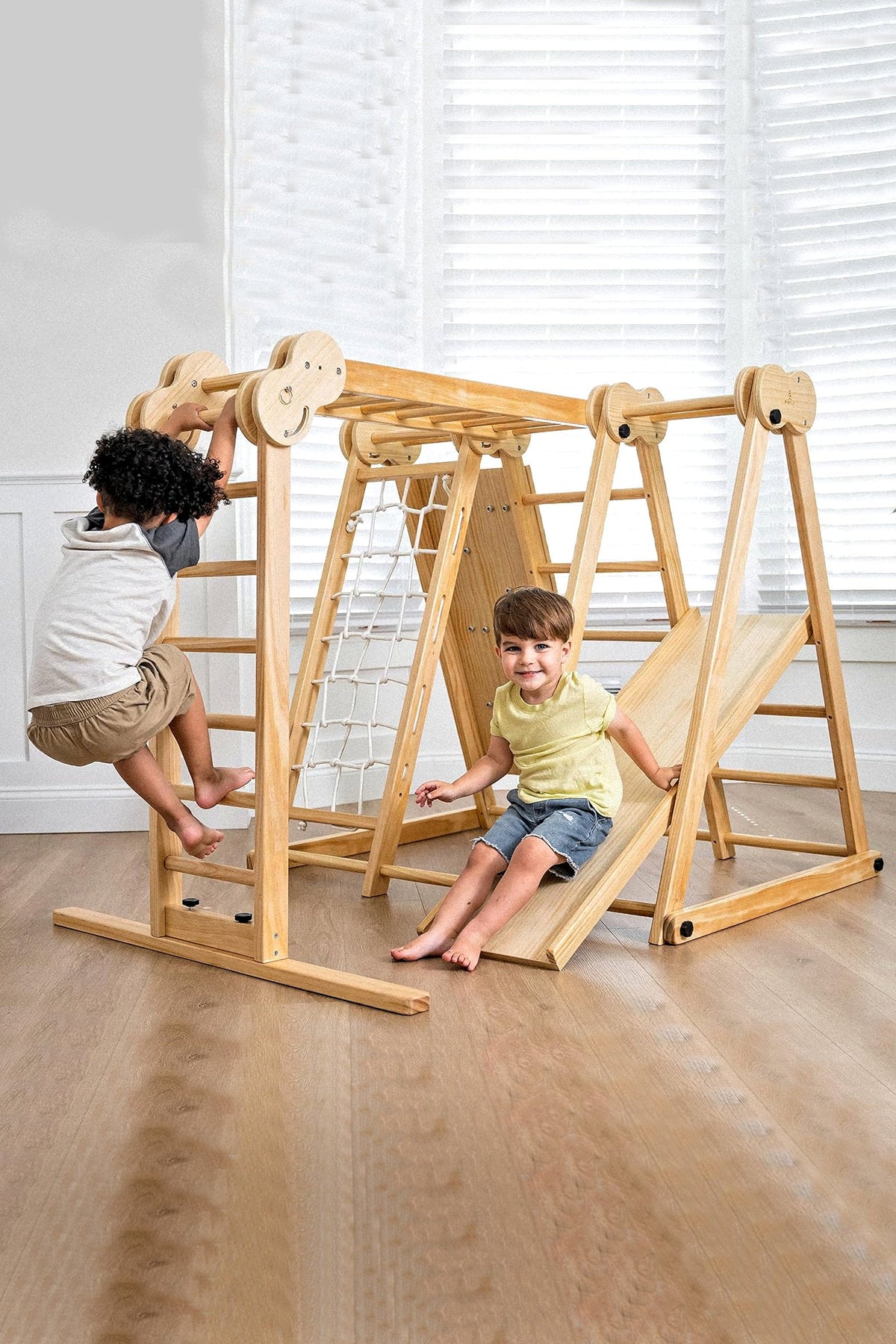 Two children play on a natural wooden jungle gym indoors, featuring climbing ladders and a slide.