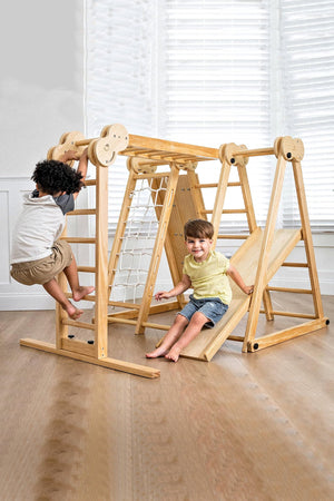 Two children play on a natural wooden jungle gym indoors, featuring climbing ladders and a slide.