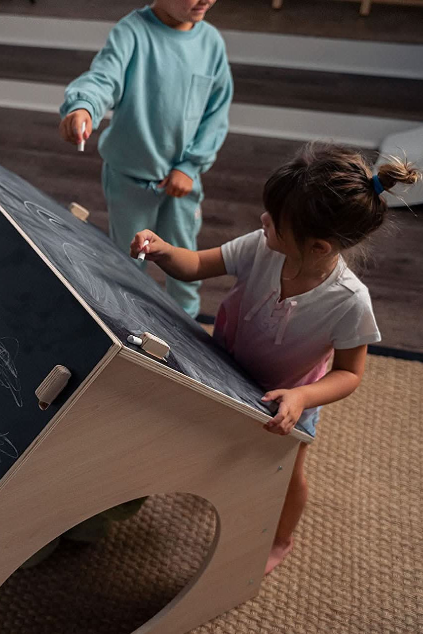 Two children engage playfully with a wooden chalkboard structure, drawing with chalk. The light wood contrasts with the dark chalk surface, creating a fun, imaginative play environment indoors.