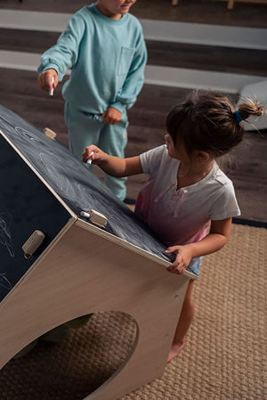 Two children engage playfully with a wooden chalkboard structure, drawing with chalk. The light wood contrasts with the dark chalk surface, creating a fun, imaginative play environment indoors.