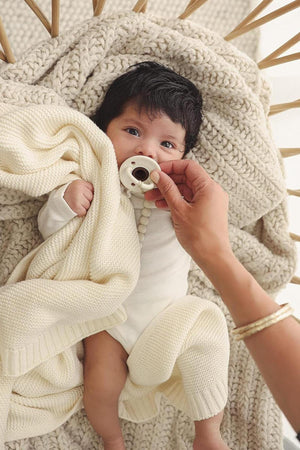 A baby wrapped in a cream knit blanket, holding a pacifier, lies on a textured knit surface.