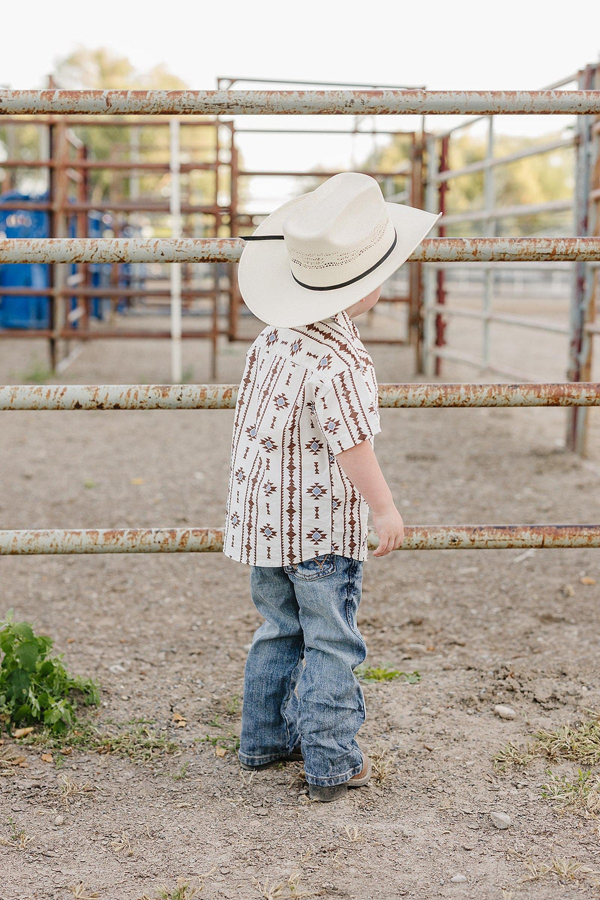 A young child wearing a white Aztec-patterned Western shirt and a wide-brimmed hat stands by a rustic metal fence. The outfit showcases a charming cowboy style perfect for adventures.