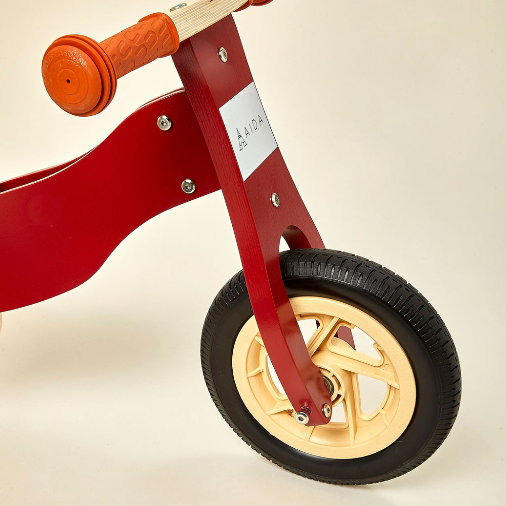 A close-up view of a red wooden balance bike showcasing its front wheel with a black tire, orange handlebar grips, and a white brand label.