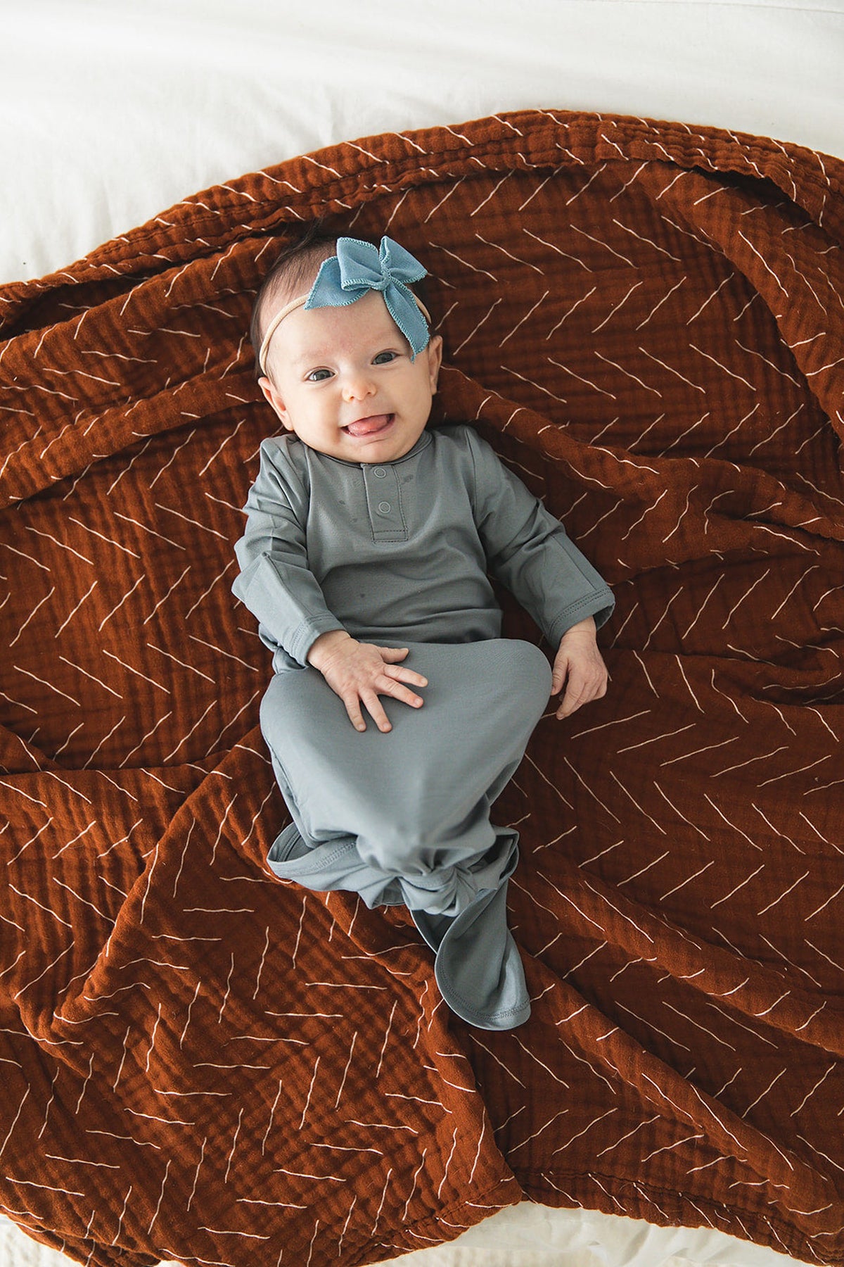 A baby wearing a dusty blue knot gown and headband lies on a patterned brown blanket, smiling.