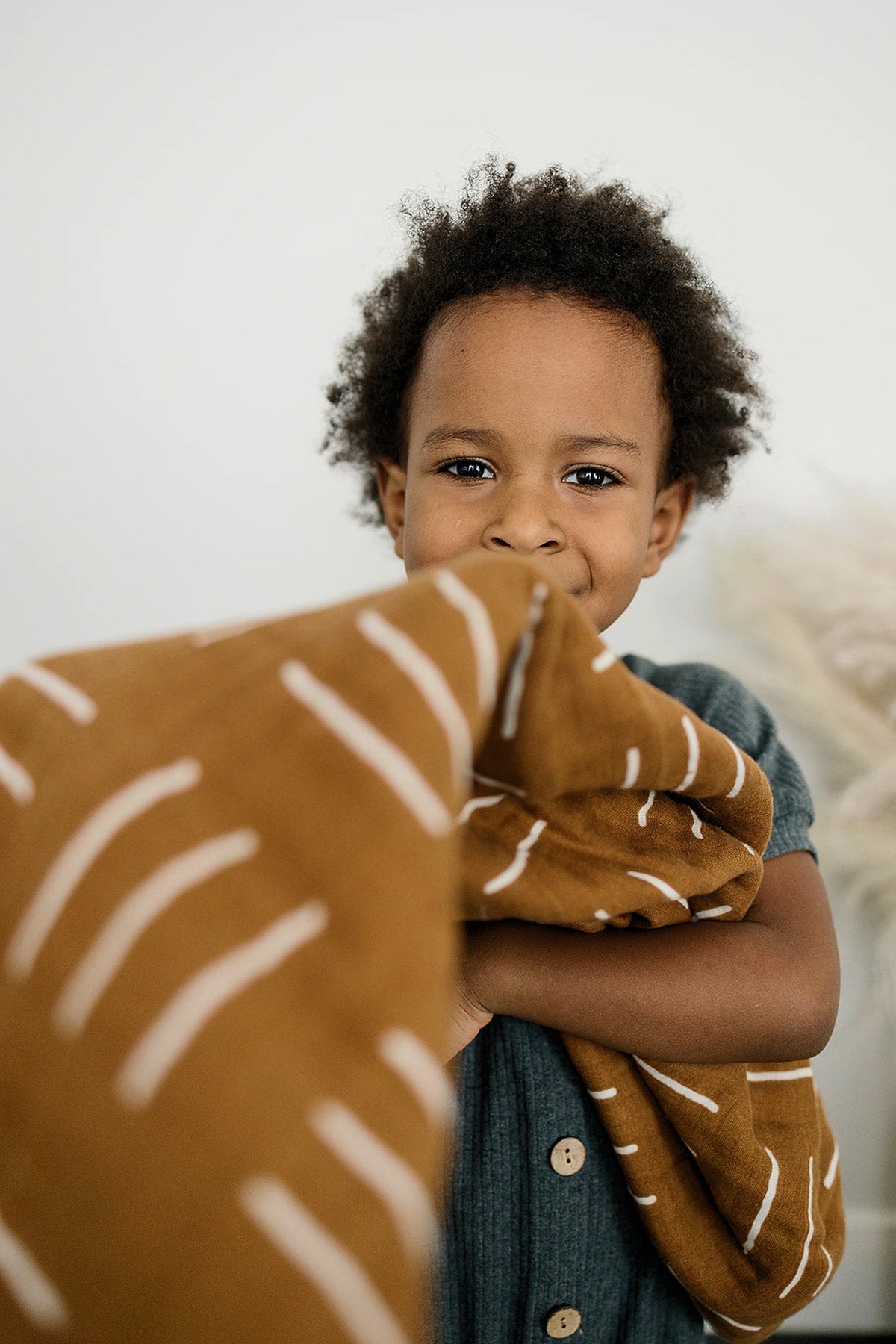 A child holds a mustard-colored quilt with white stripes, smiling gently. The quilt appears soft and cozy.