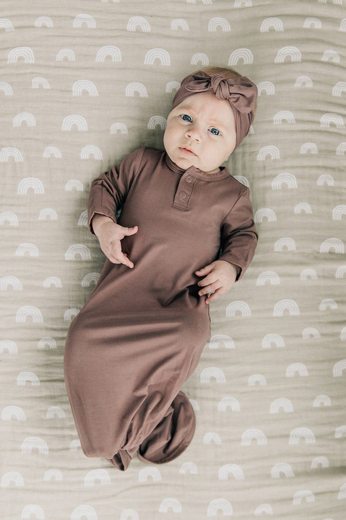 A baby wearing a plum-colored knot gown and headband lies on a patterned blanket with white rainbows.