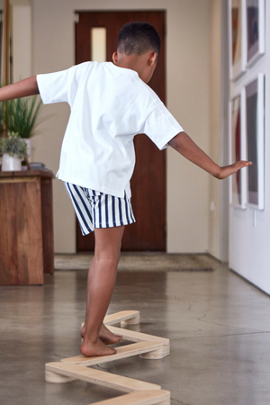A child balances on a wooden balance beam indoors, wearing a white t-shirt and striped shorts. The setting features modern decor, highlighting the beams sustainable design for active play.