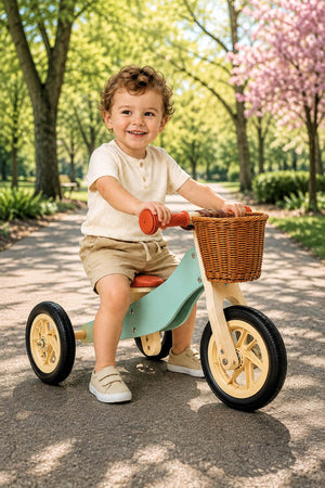 A toddler enjoys riding a mint green wooden balance bike with a wicker basket in a sunny park, surrounded by trees and blooming flowers.