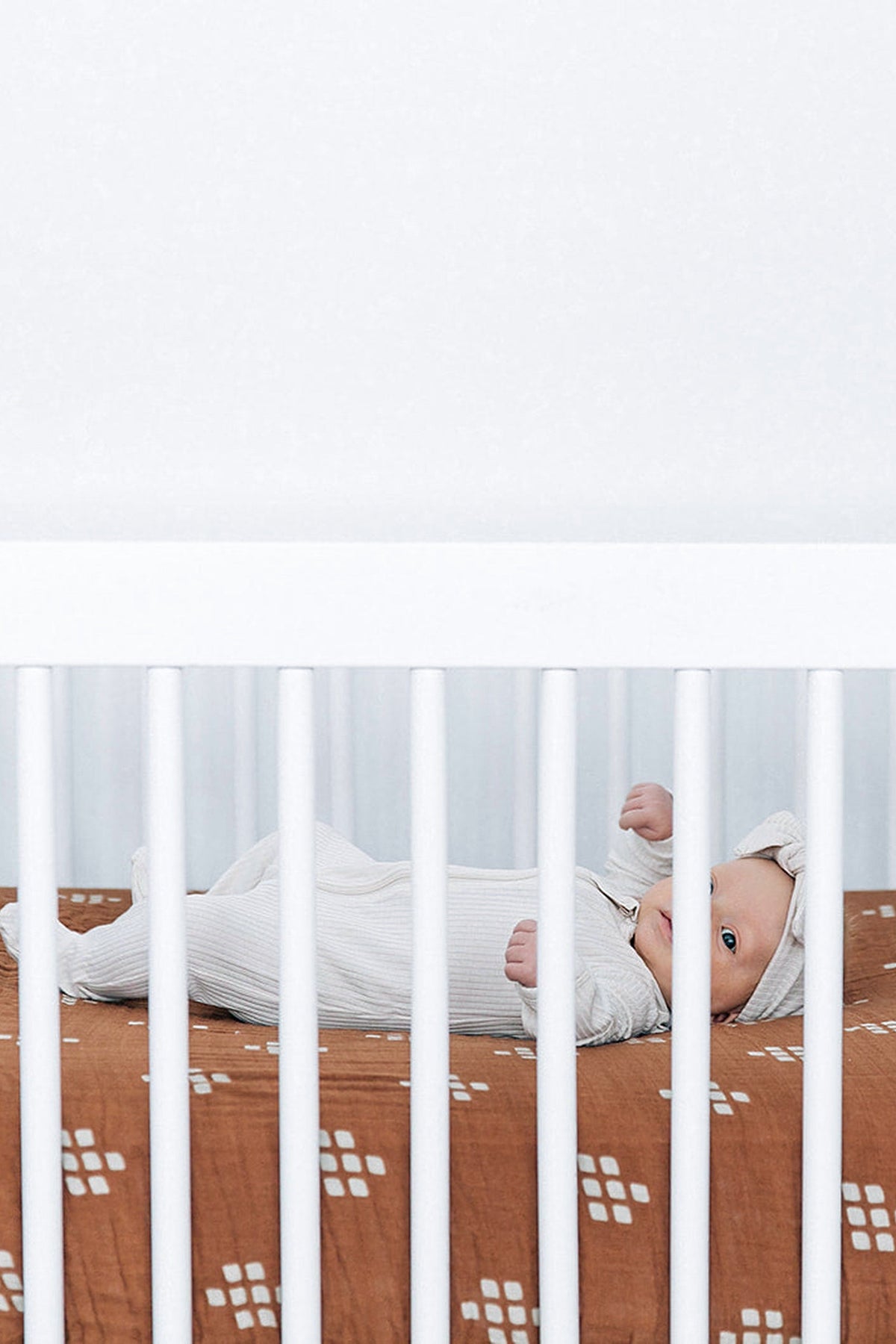 A baby lies in a crib with a chestnut muslin sheet featuring a white geometric pattern.