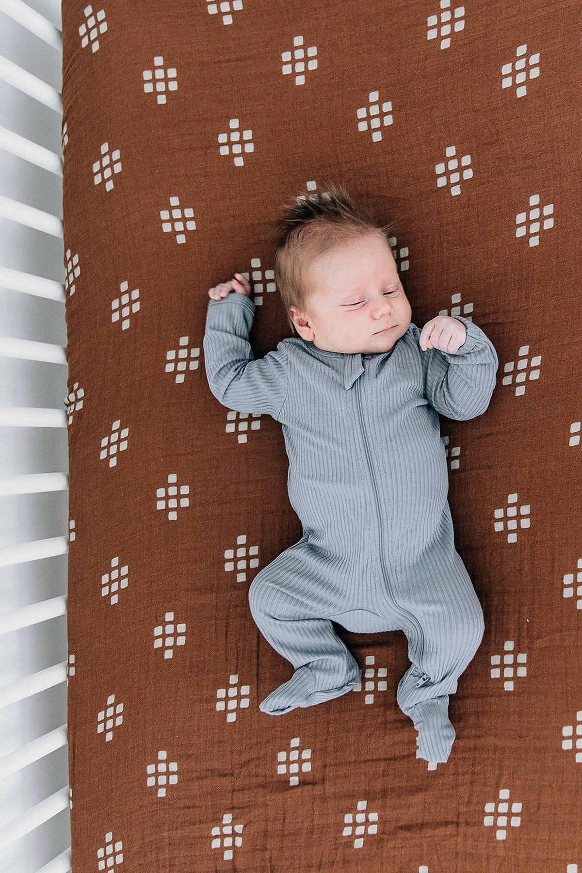 A baby sleeps on a chestnut muslin crib sheet with a geometric pattern, wearing a gray ribbed onesie.