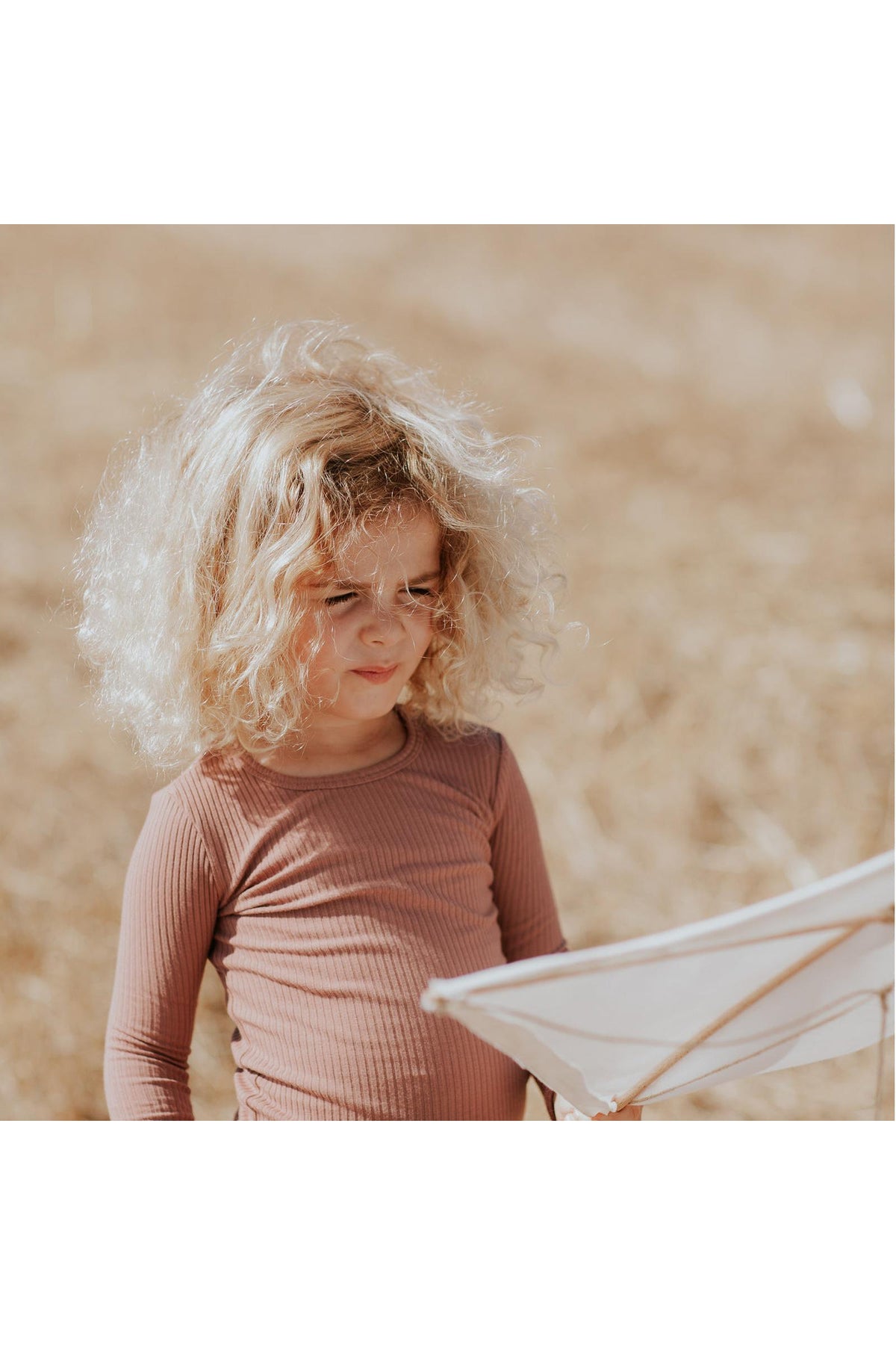 A young child with curly blonde hair stands in a field, wearing a rosewood ribbed long sleeve tee. The soft fabric complements the natural, outdoor setting and sunny atmosphere.