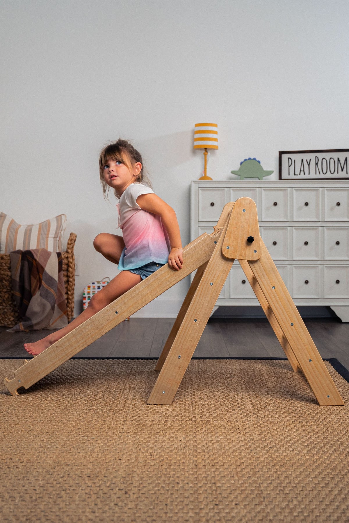 A young child climbs on a wooden Pikler triangle playset in a cozy indoor setting, featuring neutral tones and natural materials. The room includes playful decor and a woven rug.