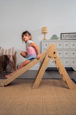 A young child climbs on a wooden Pikler triangle playset in a cozy indoor setting, featuring neutral tones and natural materials. The room includes playful decor and a woven rug.