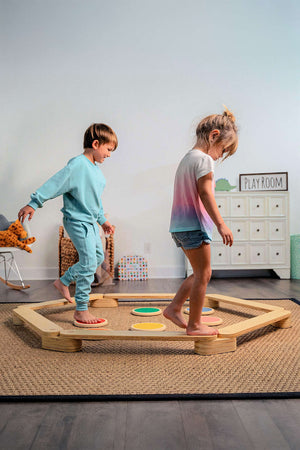 Two children play on a wooden balance beam and stepping stones set, navigating colorful circular stones on a textured rug in a bright playroom.