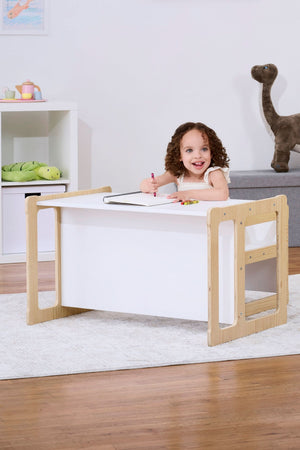 A young girl sits at a wooden toddler table with a white top, smiling as she colors. The room features a gray plush dinosaur and shelves filled with toys.