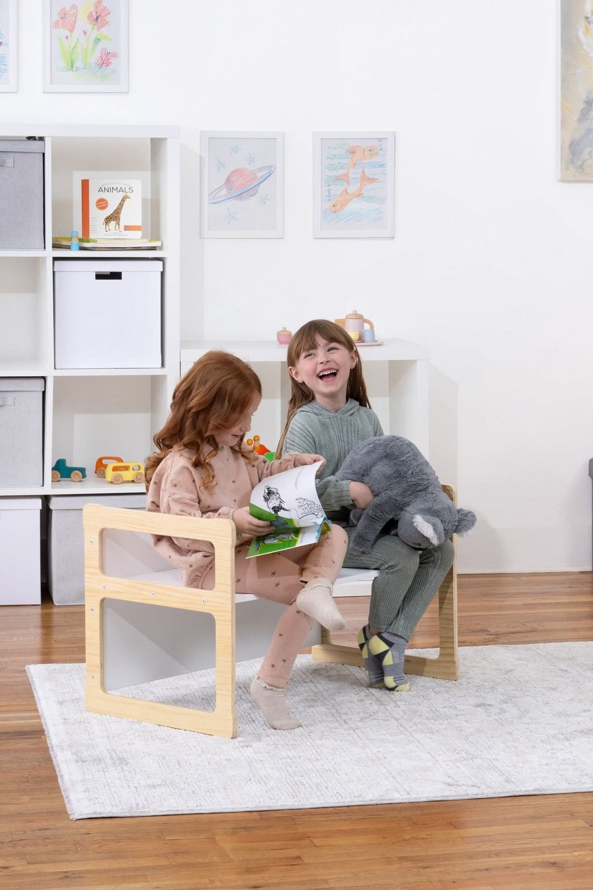 Two children sit on wooden toddler chairs, smiling and holding plush toys in a bright, modern playroom. A dinosaur plush and colorful artwork add playful elements to the space.