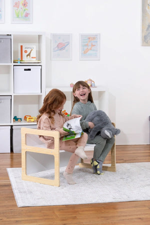 Two children sit on wooden toddler chairs, smiling and holding plush toys in a bright, modern playroom. A dinosaur plush and colorful artwork add playful elements to the space.