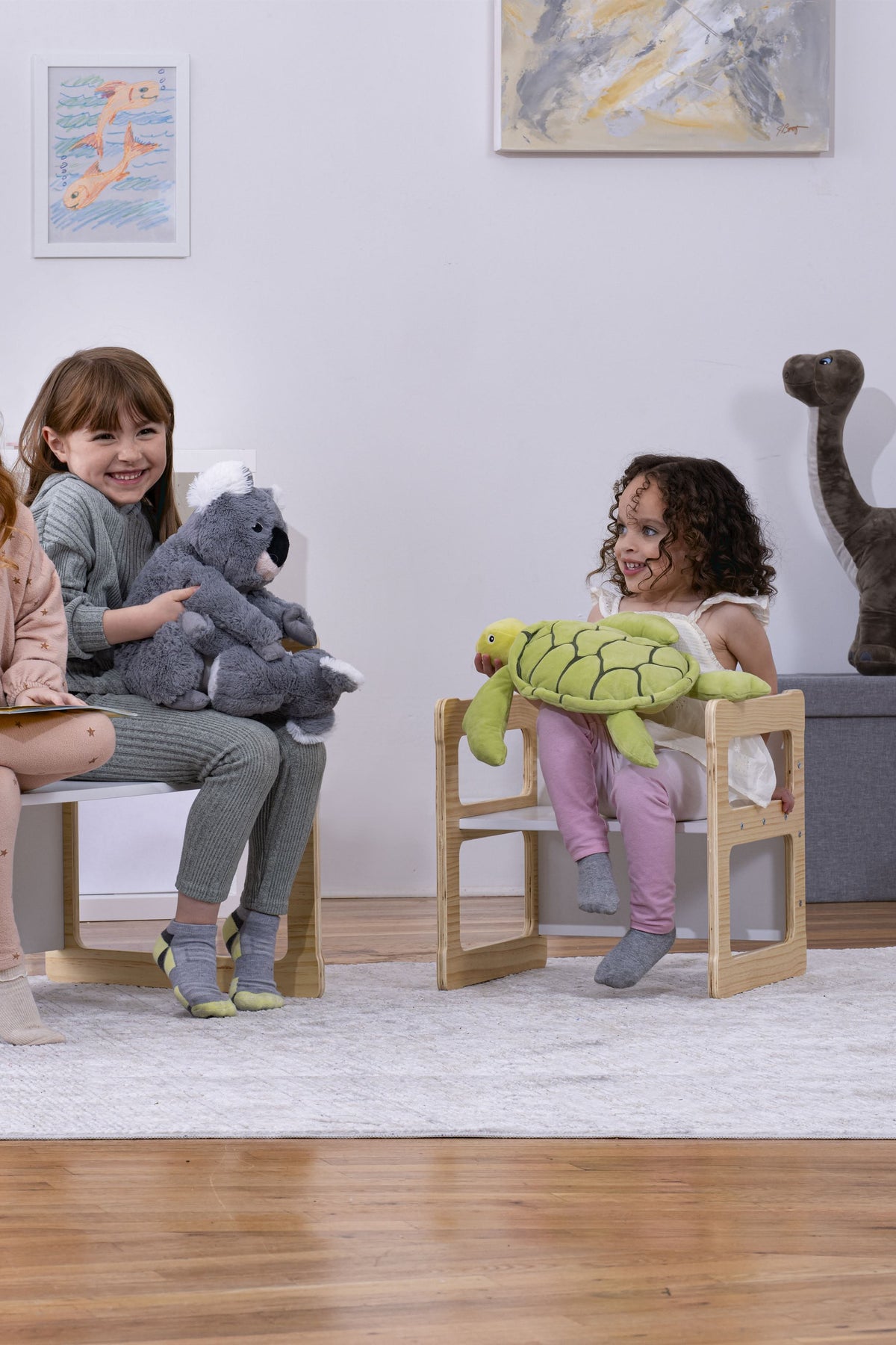 Three children sit on wooden, convertible chairs, smiling while holding plush toys. The light-colored furniture is set in a cozy, modern room with soft decor and a playful atmosphere.