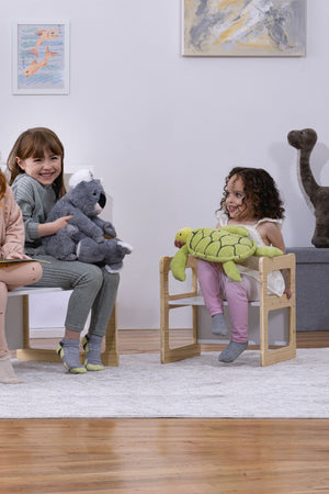 Three children sit on wooden, convertible chairs, smiling while holding plush toys. The light-colored furniture is set in a cozy, modern room with soft decor and a playful atmosphere.