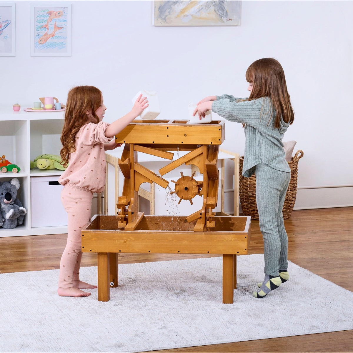 Two children play at an outdoor wooden water table, pouring water and observing its movement. The table features a natural wood finish.