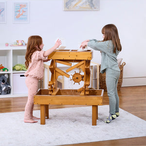 Two children play at an outdoor wooden water table, pouring water and observing its movement. The table features a natural wood finish.