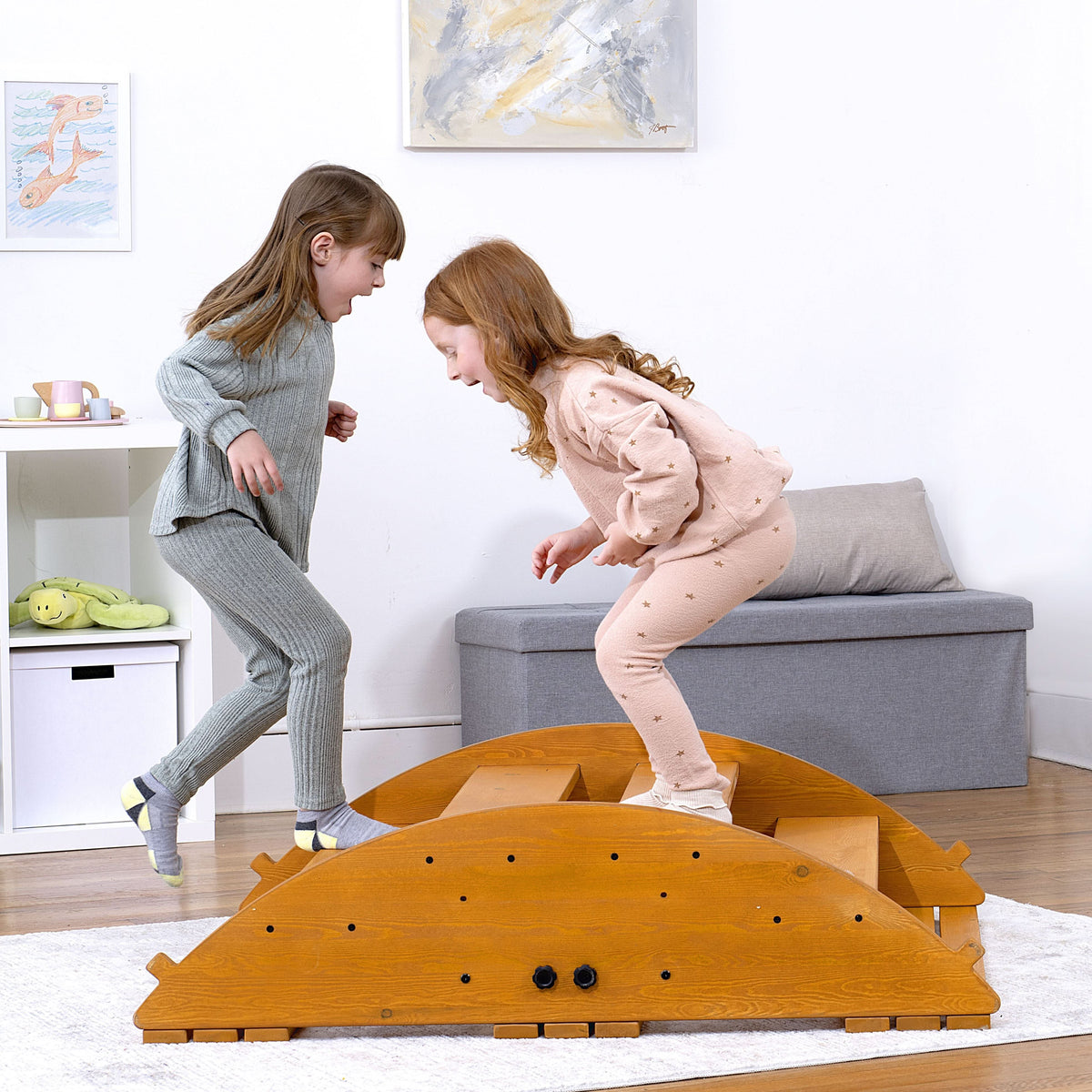 Two children joyfully play on a wooden rocker climber, smiling as they jump off its curved surface. The playset is made of sustainably sourced wood, promoting active, imaginative play.