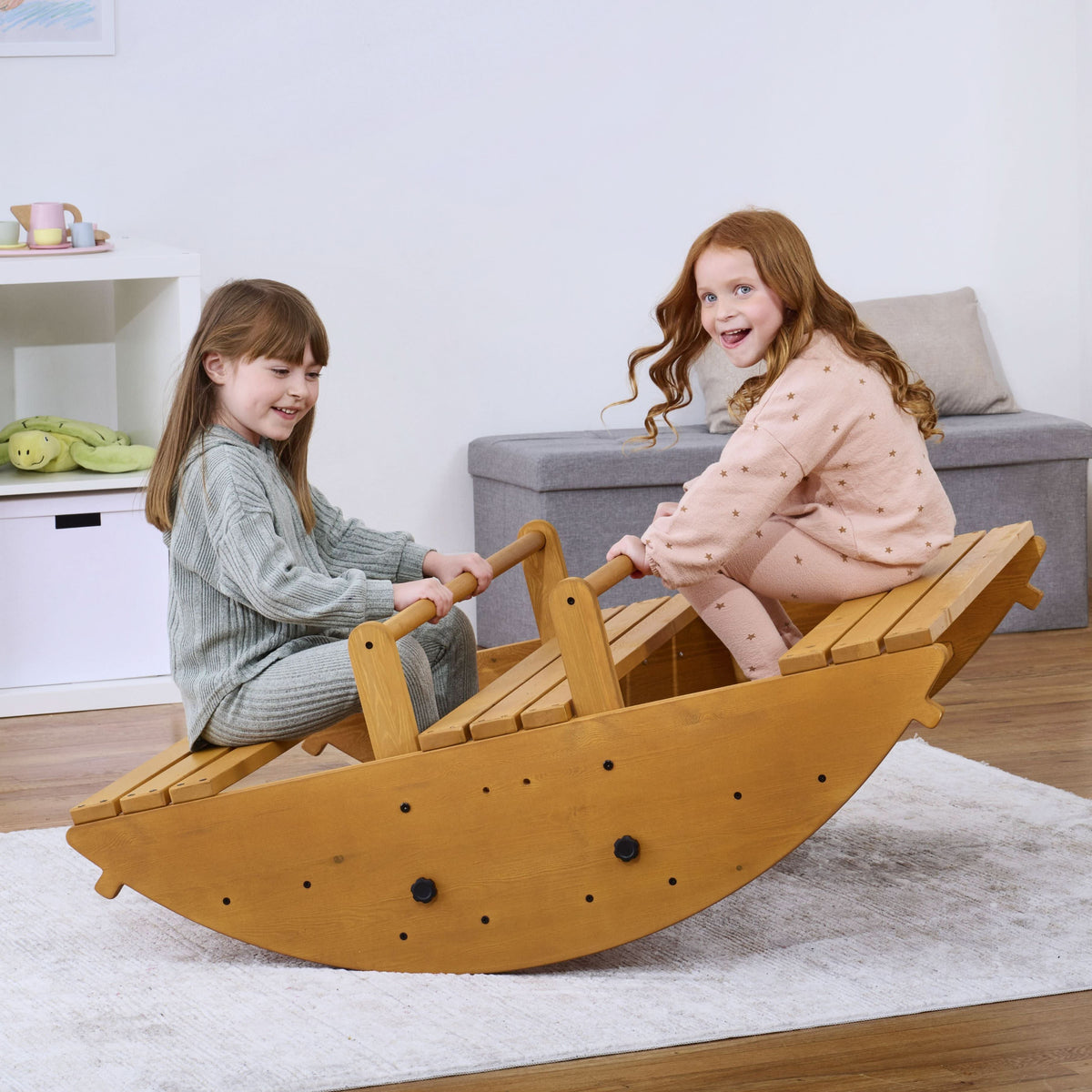 Two children joyfully play on the Banyan Jumbo Rocker, a sustainably crafted wooden climbing toy, in a bright, cozy indoor setting with light-colored flooring.