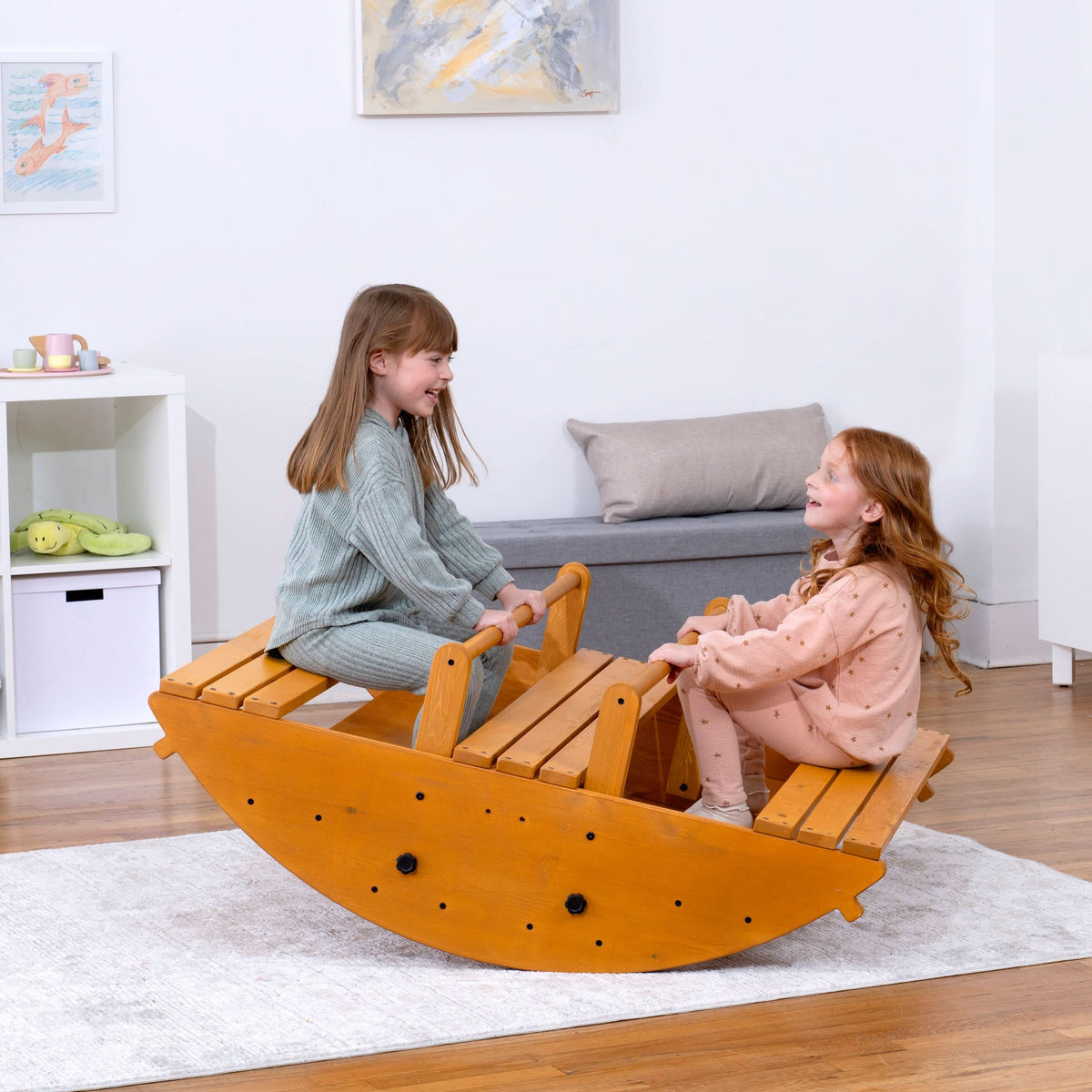 Two children sit on a large wooden rocker, smiling at each other. The toy features a natural wood finish and is set in a bright.