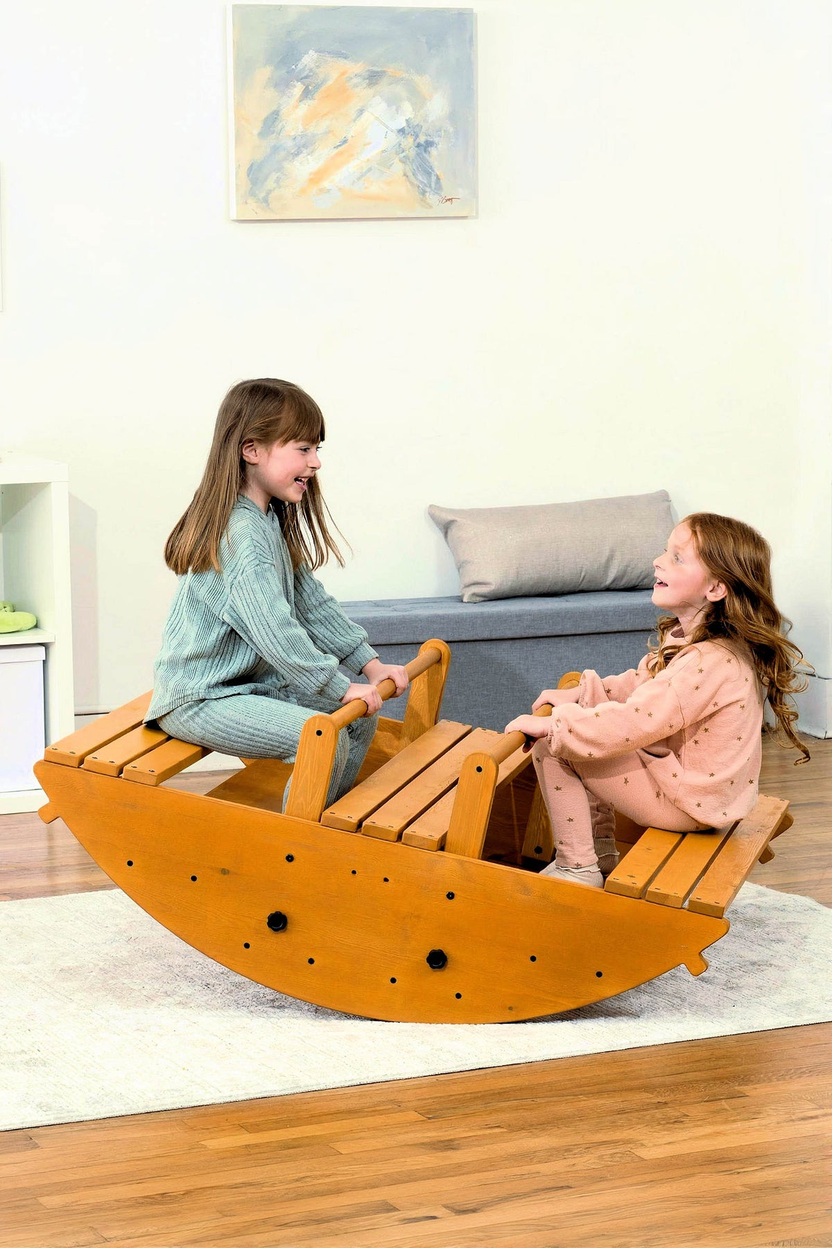Two children play on a wooden rocker, laughing together. The sturdy, natural wood construction offers a safe, engaging space for active play and balance development in a bright indoor setting.