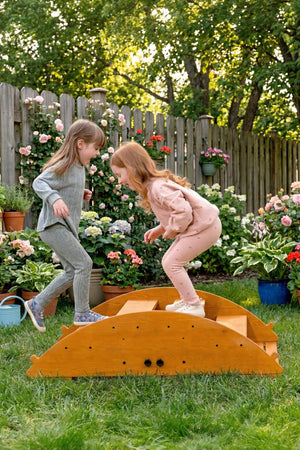 Two children play on a wooden rocker in a sunny garden, surrounded by vibrant flowers and greenery. The playset encourages active movement and balance in a natural outdoor setting.
