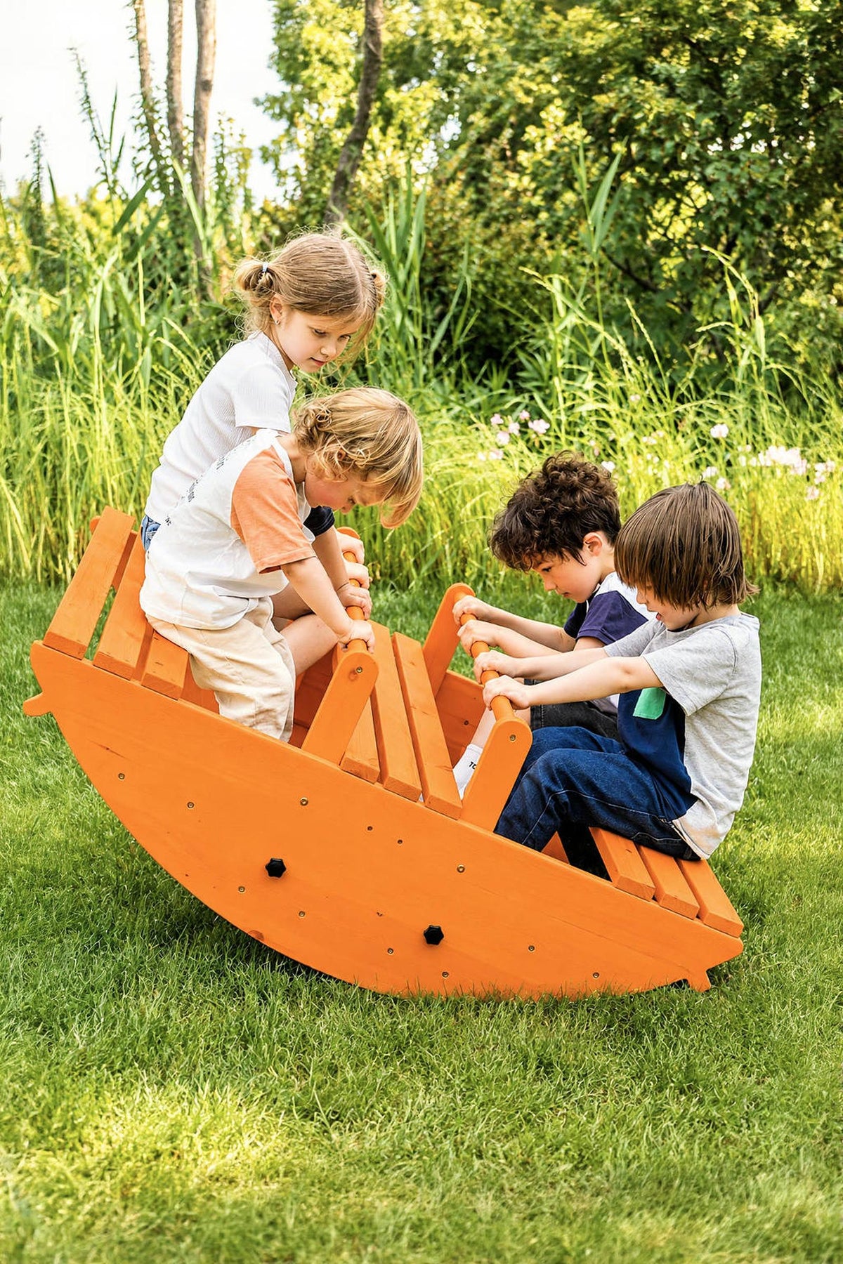 Four children play on a bright orange wooden rocker in a lush green garden. The sturdy design encourages active play and gross motor skills, making it perfect for outdoor fun.