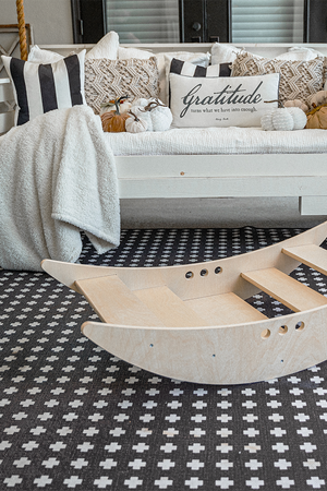 A light wooden rocking boat sits on a patterned rug, surrounded by cozy decor, including striped cushions and a swing sofa, creating a warm and inviting play space.