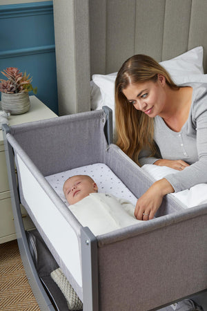 A woman gently touches a sleeping baby in a gray bedside bassinet with a breathable mattress and star-patterned sheet.