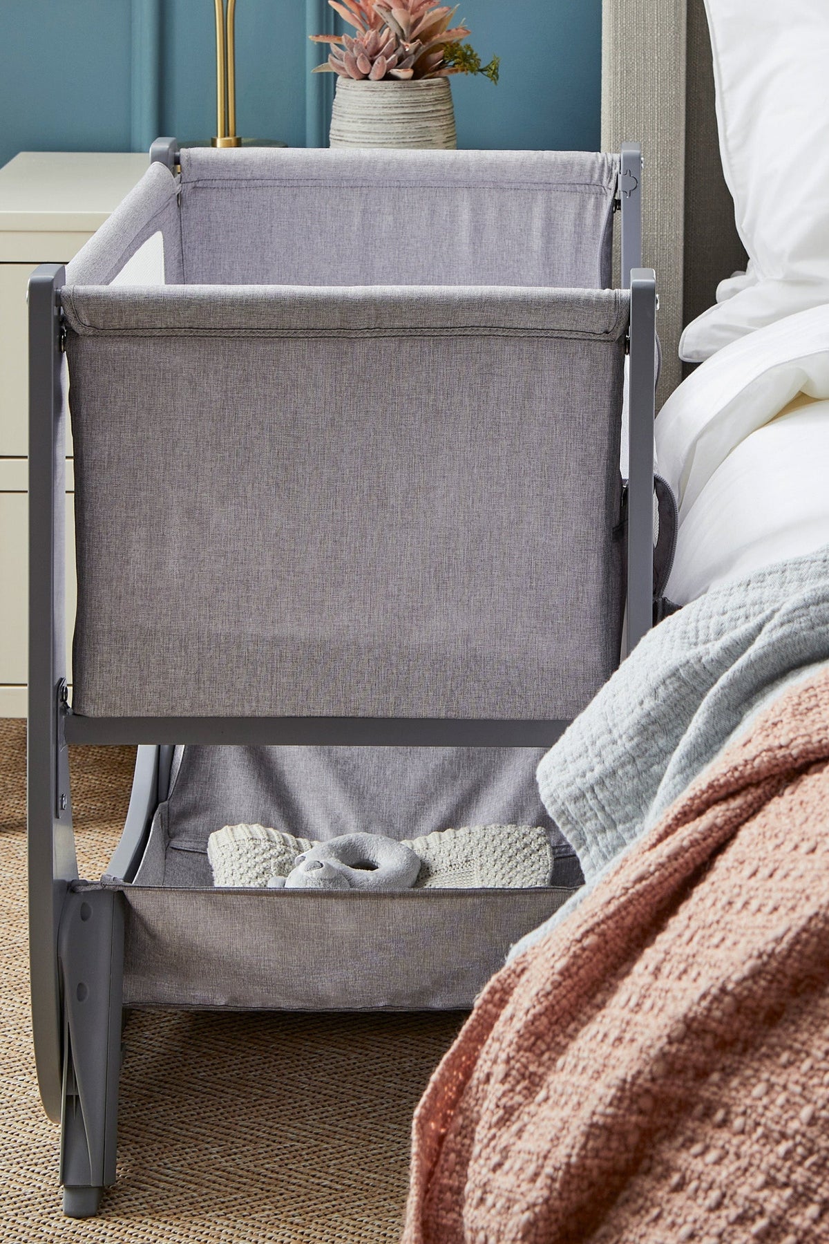A sleek, gray Shnuggle Air Lite bedside bassinet features breathable mesh sides and a height-adjustable design, positioned beside a cozy bed with a textured blanket and decorative plant nearby.