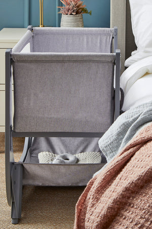 A sleek, gray Shnuggle Air Lite bedside bassinet features breathable mesh sides and a height-adjustable design, positioned beside a cozy bed with a textured blanket and decorative plant nearby.
