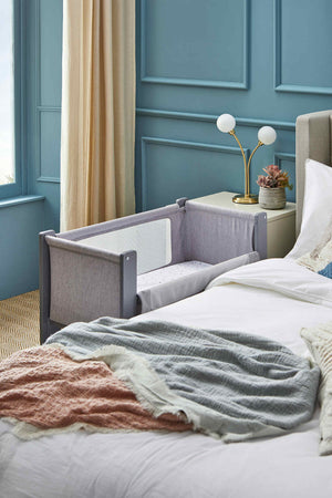 A height-adjustable Shnuggle Air Lite Bedside Bassinet in soft gray fabric features breathable mesh sides and a hypoallergenic mattress, positioned beside a cozy bed in a stylishly decorated room.