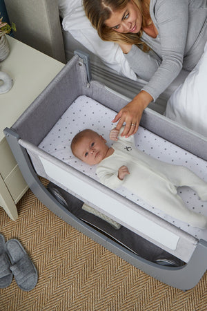 A mother gently feeds her baby lying in a light gray Shnuggle Air Lite Bedside Bassinet with breathable mesh sides.