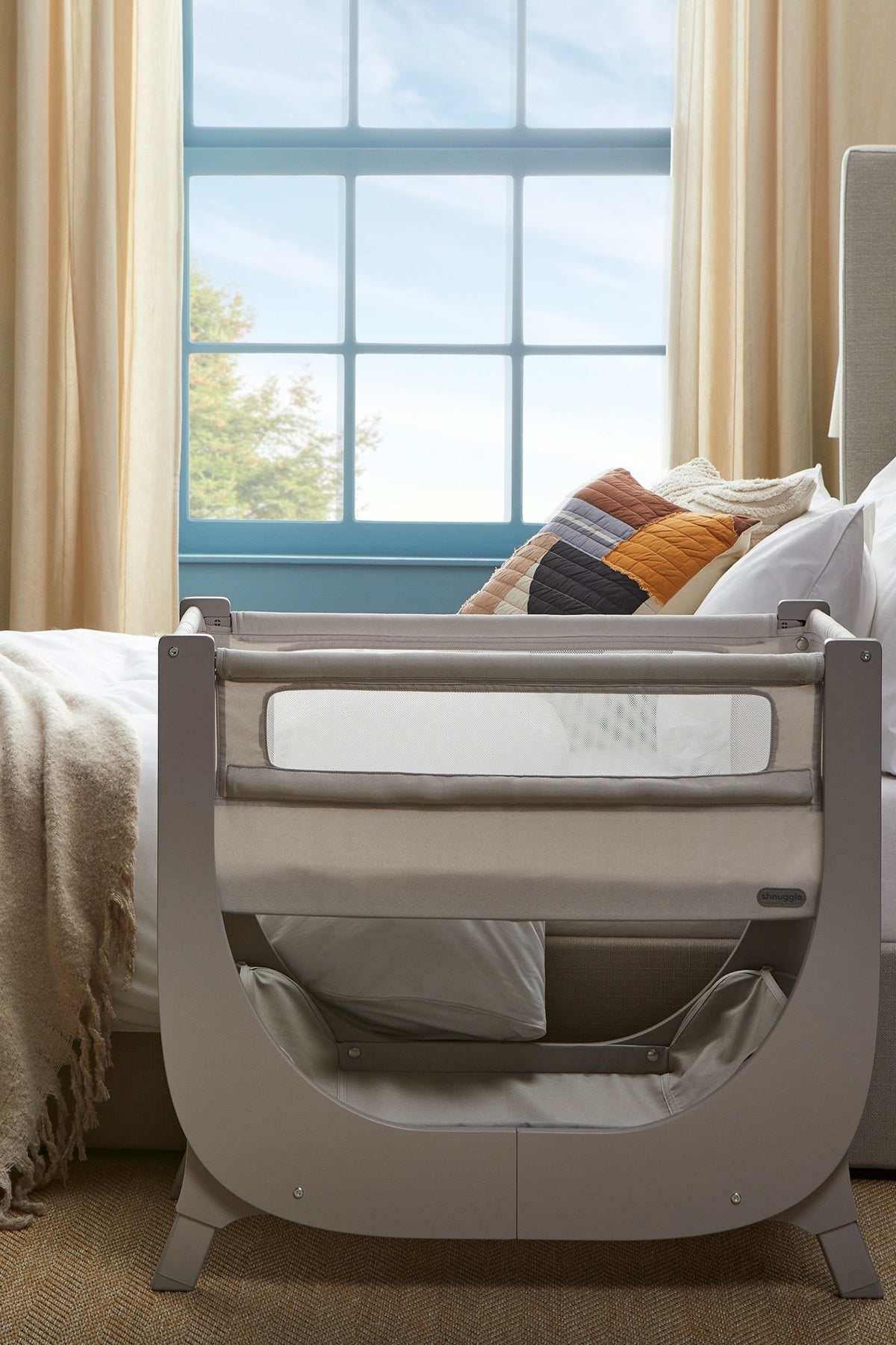 A stylish Shnuggle Air Lite bedside bassinet features breathable mesh sides and a sleek, gray wooden frame. It is positioned next to a bed with cozy pillows and soft curtains.