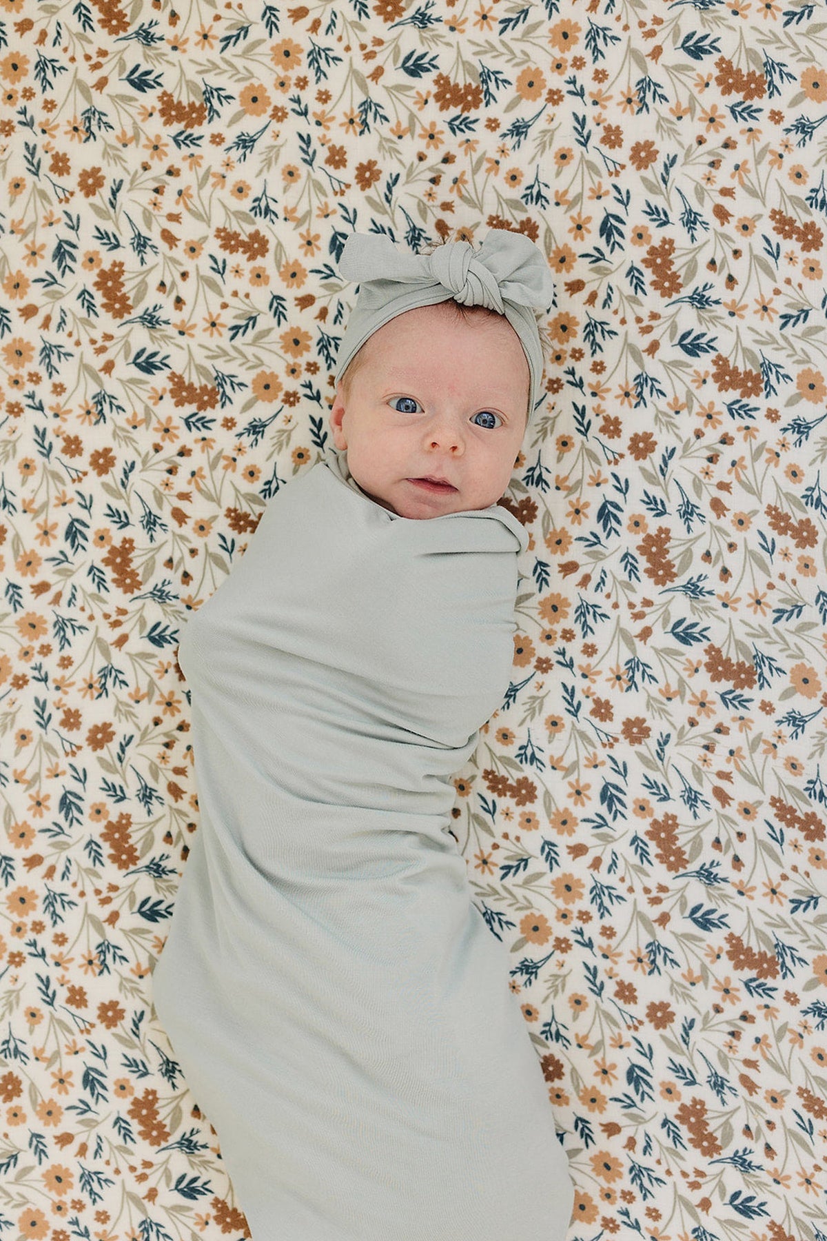 A baby in a sage green swaddle and matching headband lies on a floral-patterned blanket.