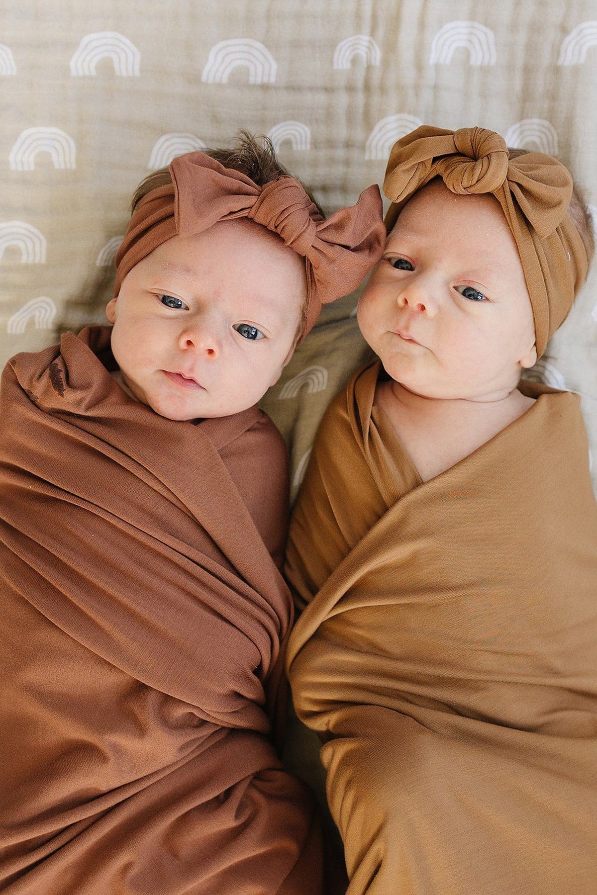 Two babies in rust and brown swaddles with matching head wraps, lying on a patterned blanket.