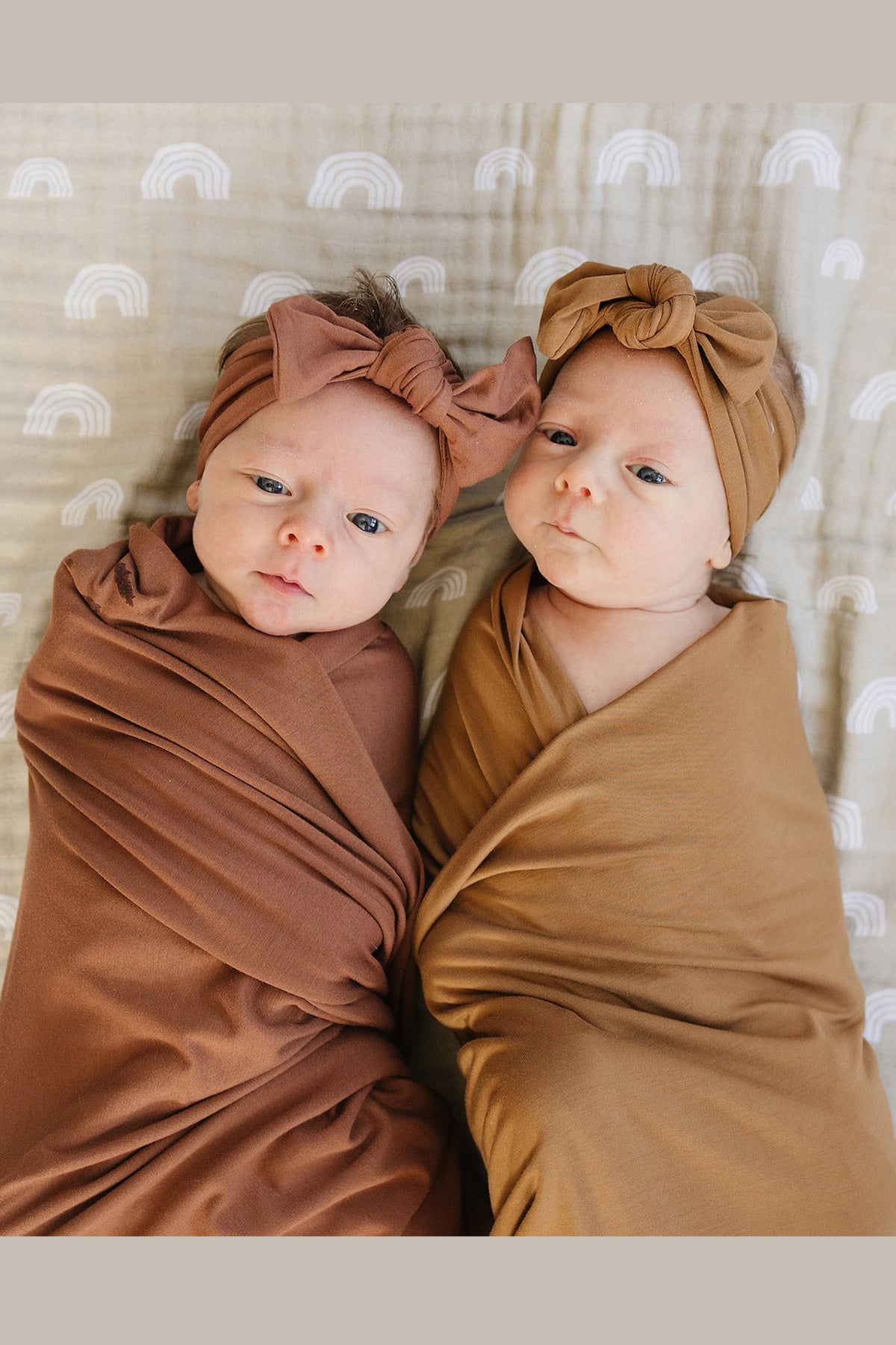 Two babies swaddled in brown blankets with matching headbands lie on a patterned blanket with white rainbows.