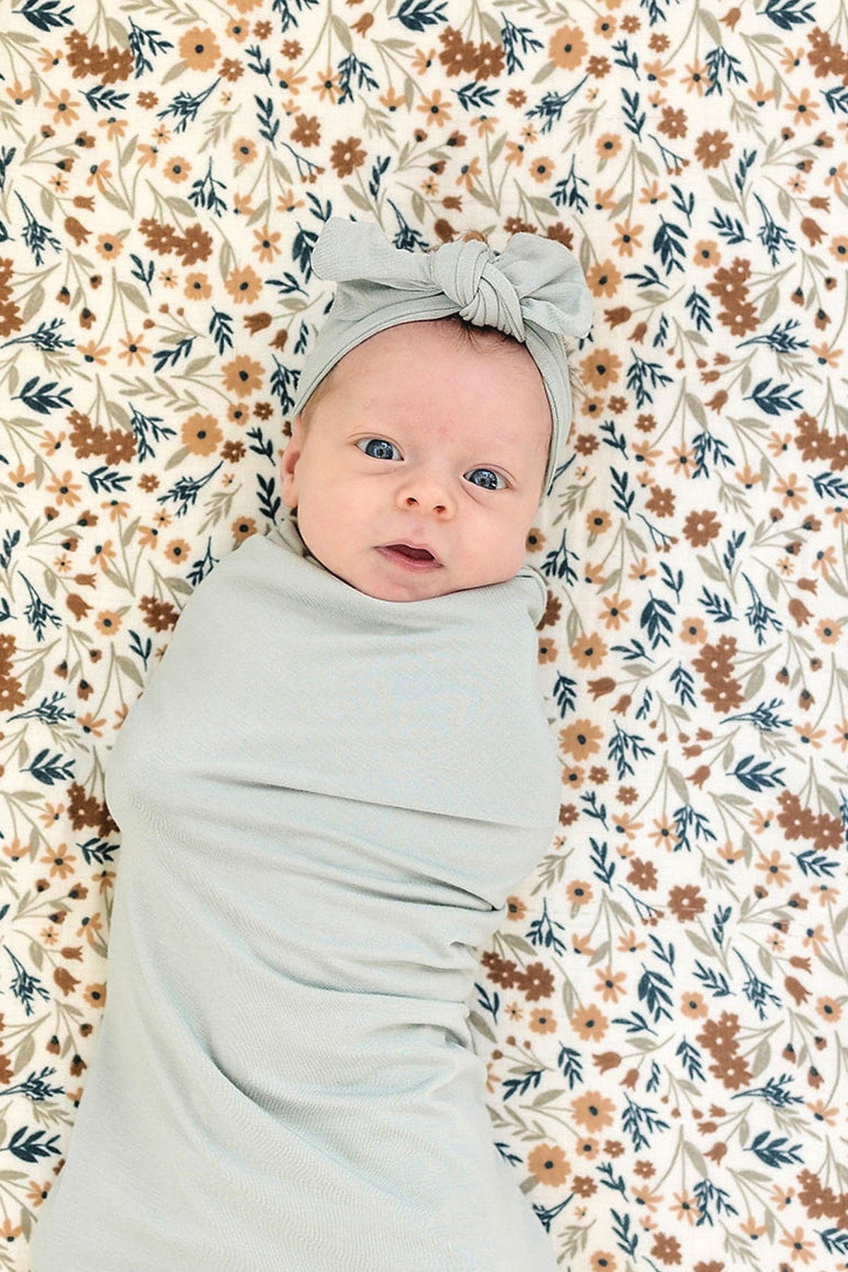 Baby wearing a sage head wrap and swaddle on a floral-patterned blanket, showcasing soft bamboo fabric.