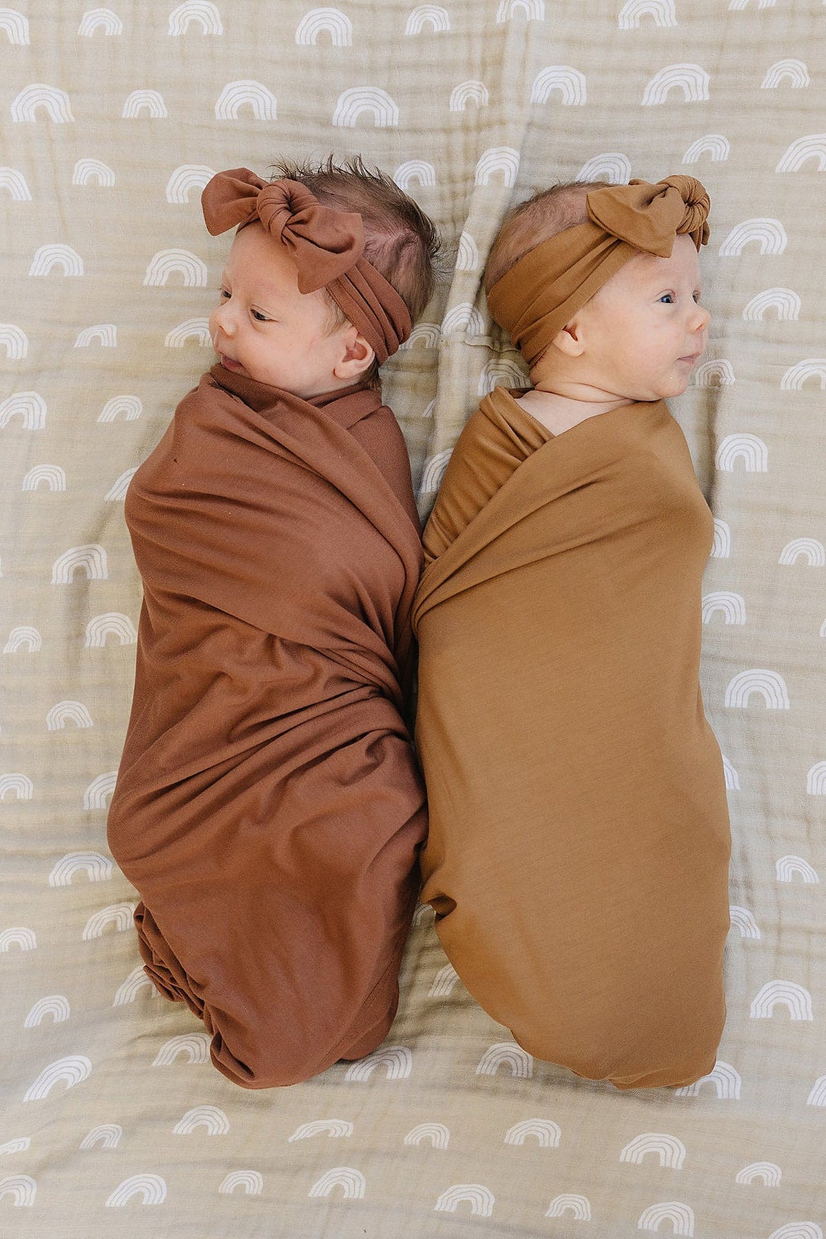 Two babies wrapped in rust-colored swaddle blankets with matching headbands lie on a patterned blanket.