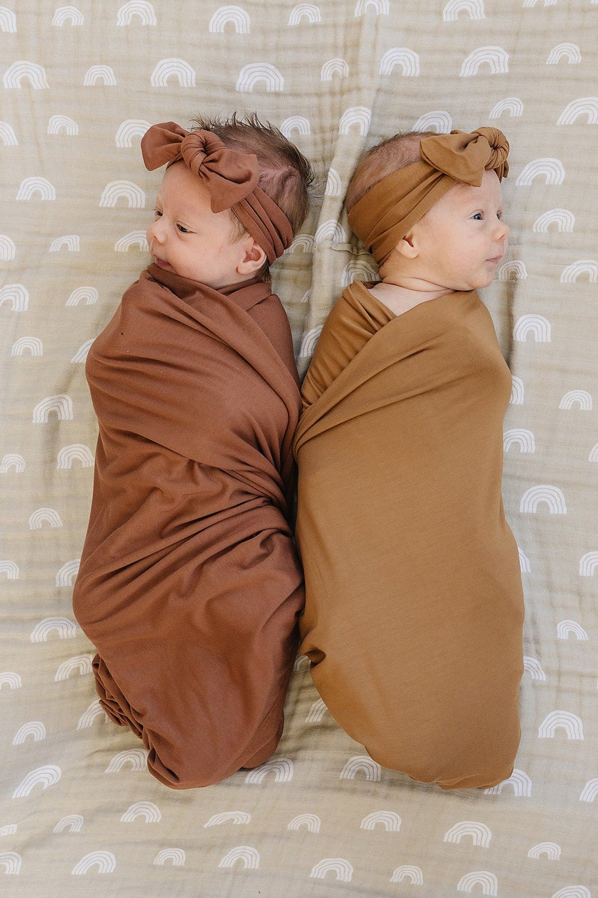 Two babies wrapped in brown bamboo swaddles with matching headbands, lying on a patterned blanket.