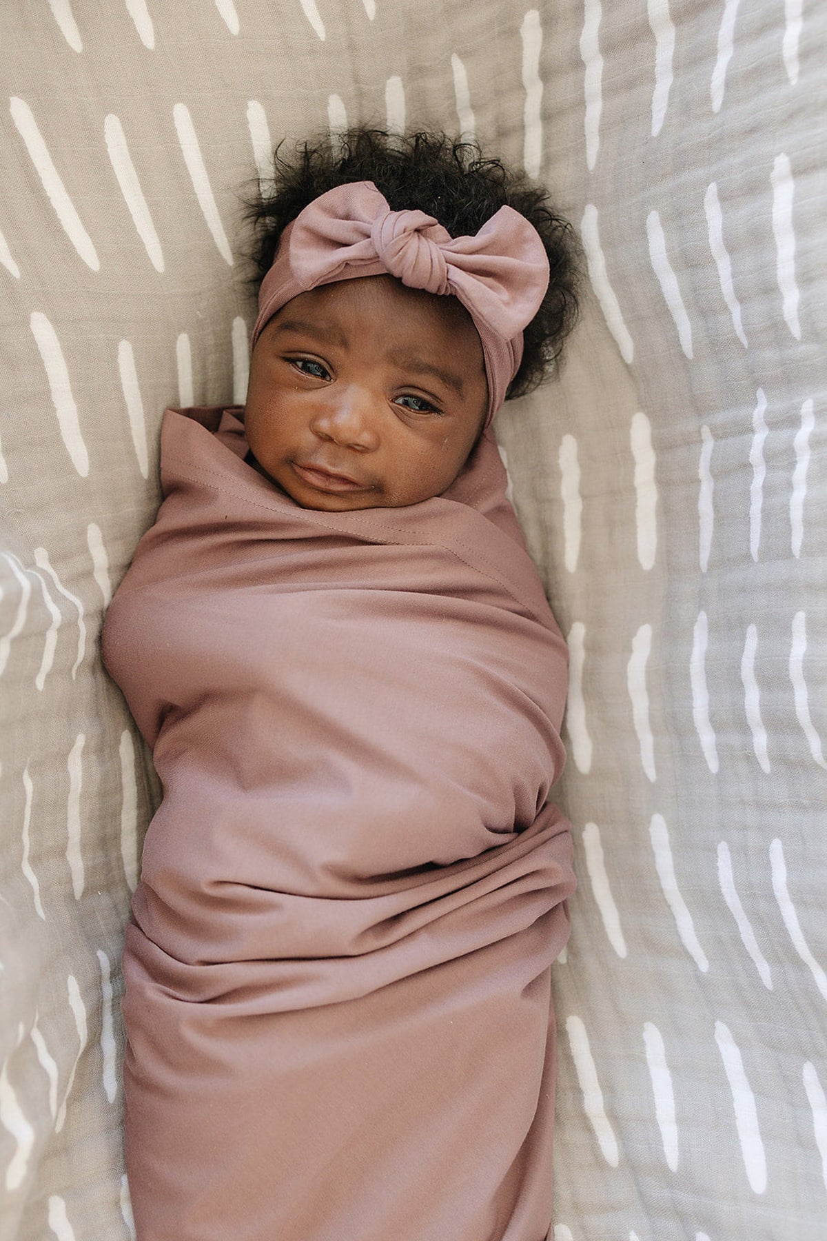 A baby wrapped in a dusty rose bamboo blanket with a matching head wrap, lying on a patterned fabric.
